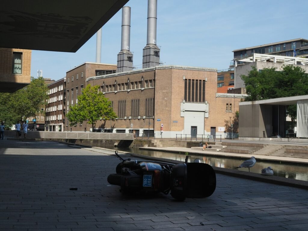 A fallen scooter on a paved canal side with industrial chimneys in the background